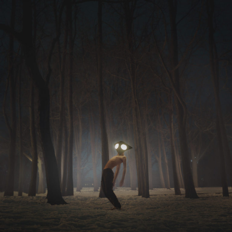 In einem nächtlichen, kahlen Wald steht eine Figur mit bloßem Oberkörper, deren Kopf durch eine leuchtende Projektorlampe ersetzt wurde – surreal und verstörend schön.