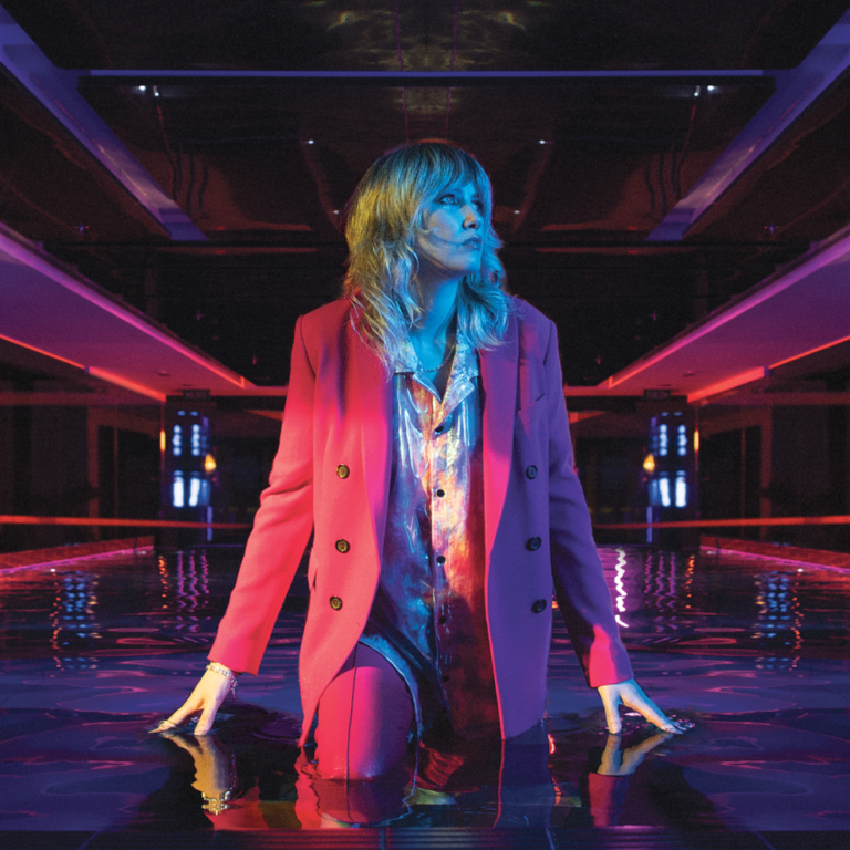 Ladyhawke kniet im pinken Anzug auf spiegelndem Boden unter Neonlicht – zwischen Club-Ästhetik und selbstbewusster Zurückhaltung.