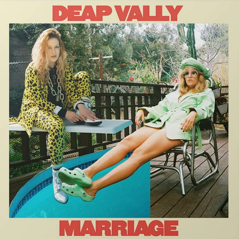 Zwei Musikerinnen von Deap Vally sitzen lässig am Pool – die eine im Leo-Anzug auf dem Sprungbrett, die andere in grünem Mantel und Crocs – zwischen Glamour und Gelassenheit.