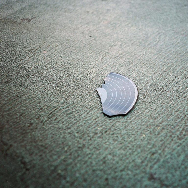 Ein zerbrochenes Stück einer Schallplatte liegt auf grünlich strukturierter Fläche – minimalistisches Cover zum Album BOOK von They Might Be Giants.