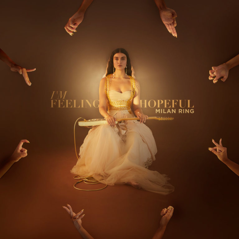 Milan Ring sitzt in weißem Tüllkleid mit Gitarre im Schoß, umgeben von symbolischen Händen – Anlehnung an die Göttin Saraswati.
