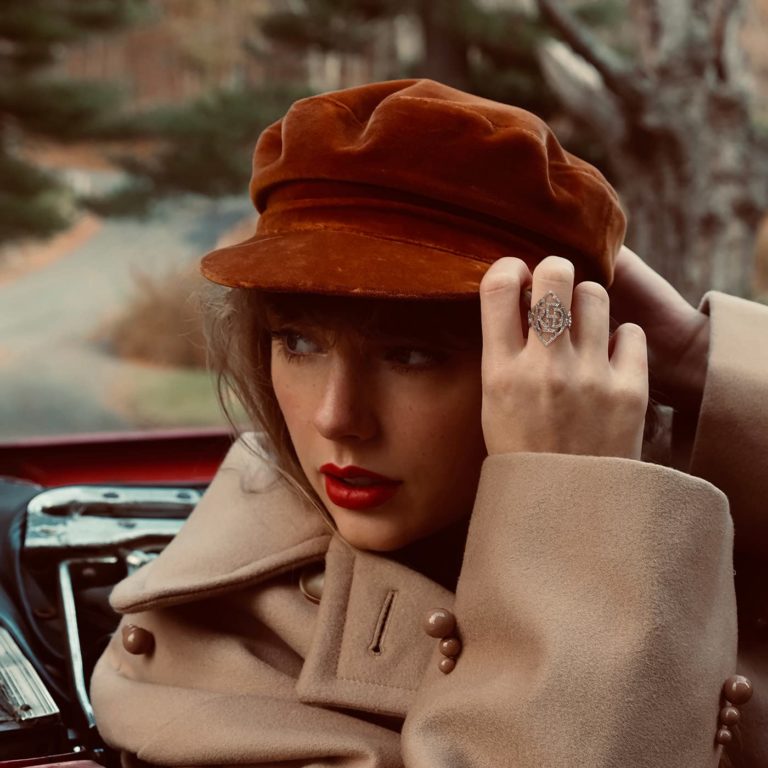 Taylor Swift sitzt mit rotem Lippenstift, beigefarbenem Mantel und rostbrauner Schirmmütze im Cabrio – nostalgisch und elegant inszeniert, mit Fokus auf Herbstästhetik.