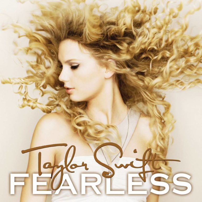 Taylor Swift mit wehenden Locken vor hellem Hintergrund. Sie trägt ein weißes Top und blickt seitlich mit geschlossenen Lippen aus dem Bild.