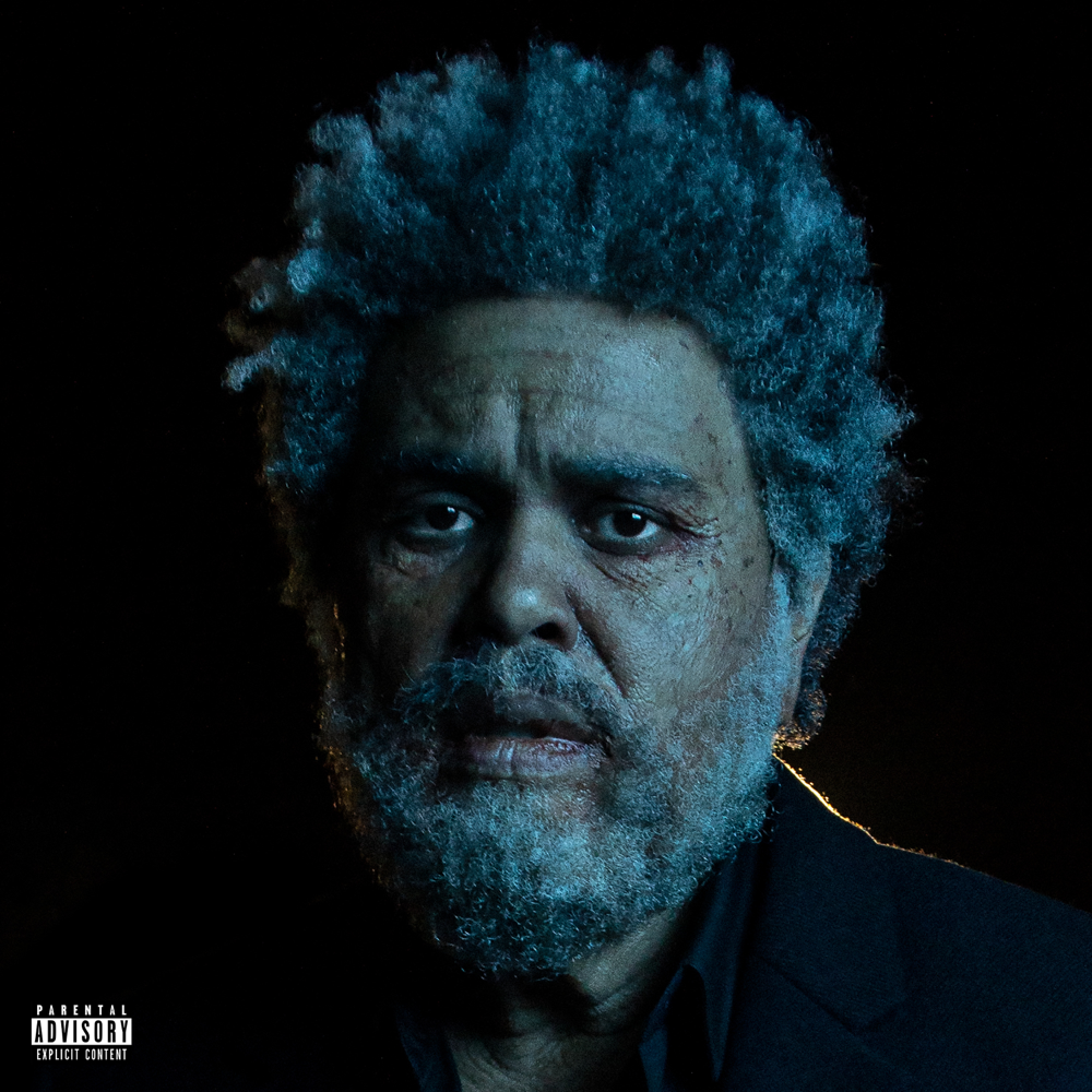 Porträt von The Weeknd mit grauen, buschigen Haaren und Bart auf schwarzem Hintergrund, trägt dunkles Sakko und wirkt nachdenklich.