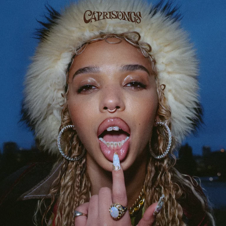 Nahaufnahme von FKA twigs mit flauschiger Pelzmütze, großen Creolen und herausgestreckter Zunge, auf der ein verziertes Zahn-Bling glänzt, vor einer Dämmerungs-Stadtsilhouette.