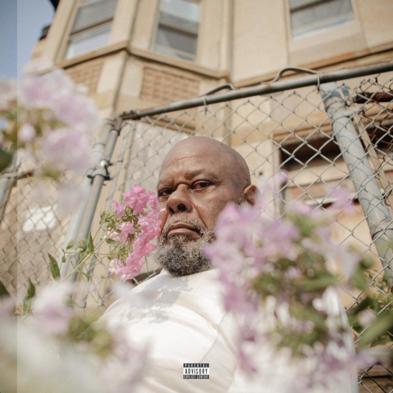 Älterer Afroamerikaner mit kahlem Kopf und grauem Bart im weißen Shirt, zwischen rosa Phlox-Blüten vor Maschendrahtzaun und Sandsteinhaus – Albumcover Few Good Things von Saba