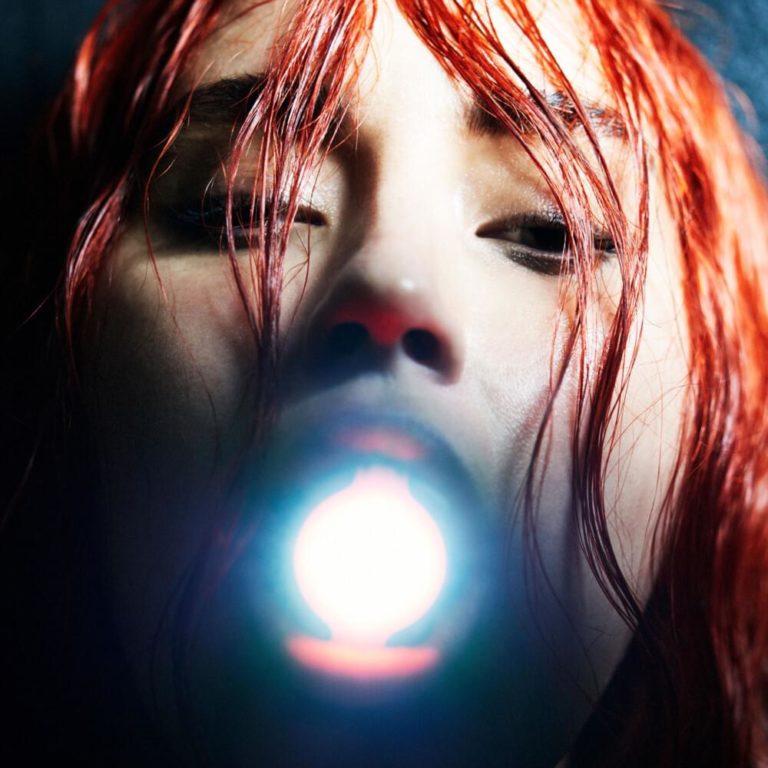 Cover des Albums „Mother“ von Highasakite – eine Nahaufnahme eines Gesichts mit roten Haaren, das von unten beleuchtet wird; der Mund öffnet sich zu einem grellen Licht, das aus dem Inneren zu strahlen scheint.