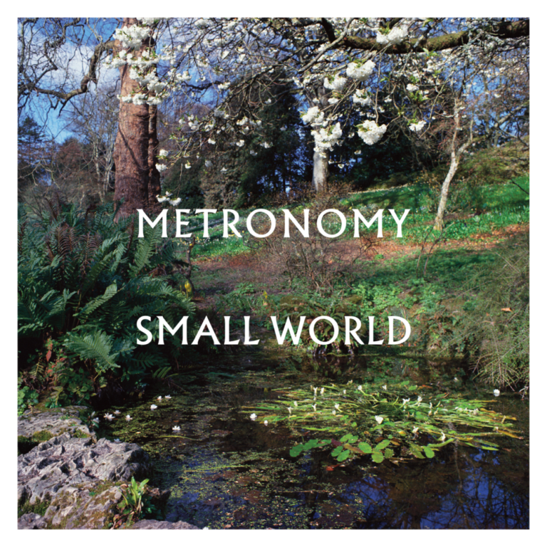 Cover des Albums „Small World“ von Metronomy – ein idyllischer Park mit Teich, Frühblühern, blühenden Bäumen und stillem, reflektierendem Wasser in malerischer Natur.