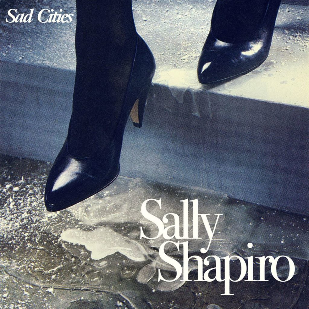 Nahaufnahme schwarzer High Heels auf einer nassen, glänzenden Treppe – das Cover zu Sally Shapiros „Sad Cities“ in nostalgisch-körniger Fotografie.