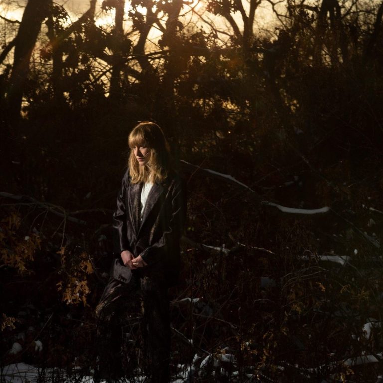 Tamara Lindeman steht in einem dunklen, winterlichen Waldstück, von goldenem Licht durchflutet, als sei sie selbst eine melancholische Lichtquelle.