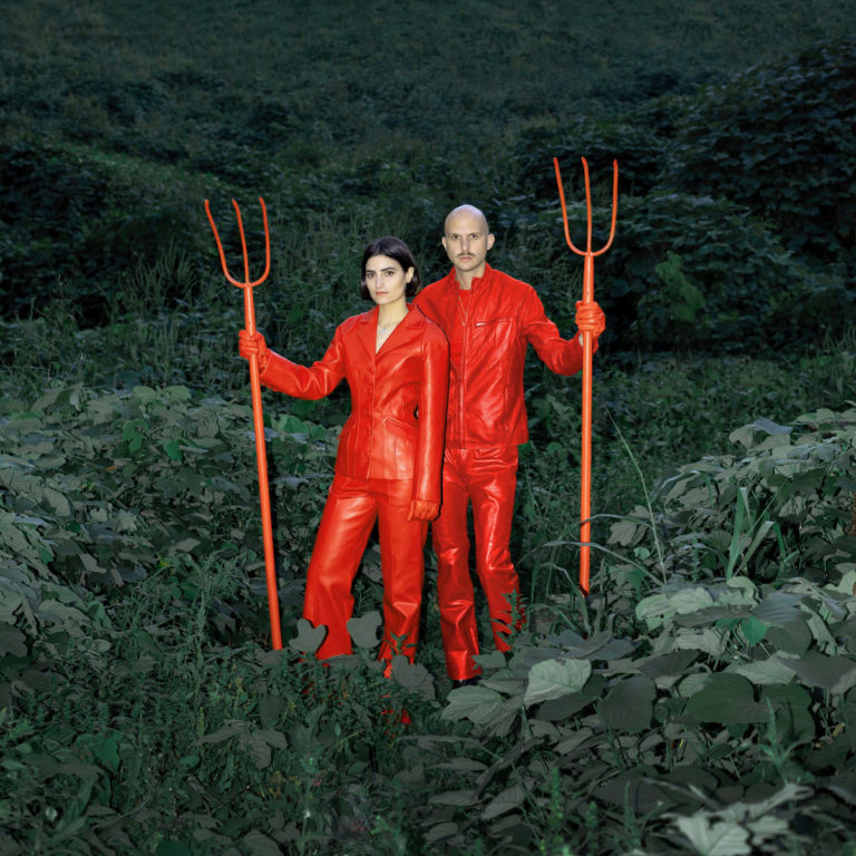 Mattiel Brown und Jonah Swilley stehen in leuchtend roten Lederjacken und -hosen im dichten, grünen Unterholz und halten jeweils eine rote Heugabel.
