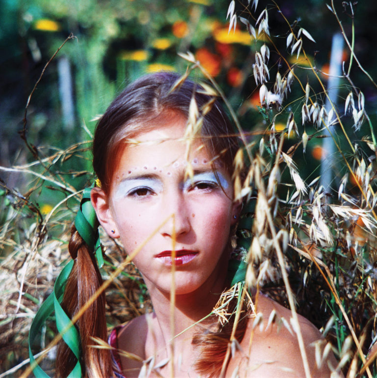 Nahaufnahme von Maia Friedman mit hellblauem Augen-Make-up und grünen Haarbändern, umgeben von goldenen Ähren und verschwommenen Wildblumen im Hintergrund.