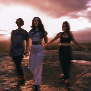 Drei verschwommene Gestalten, die im warmen Licht der untergehenden Sonne über felsiges Terrain schreiten, eingefangen in einem flüchtigen Moment der Bewegung.