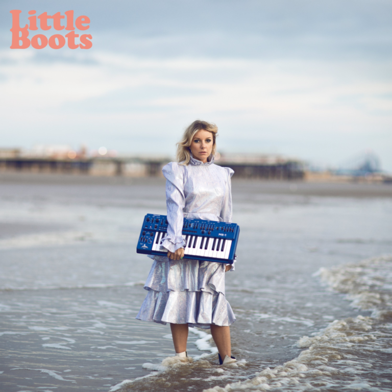 Victoria Hesketh alias Little Boots steht im seichten Meerwasser mit einem blauen Keyboard, hinter ihr Pier und endloser Himmel.