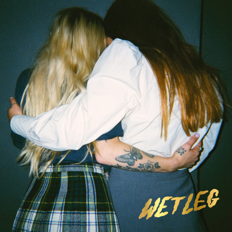 Zwei Frauen umarmen sich von hinten, eine trägt einen karierten Rock, die andere Tattoos am Arm, beide stehen vor dunklem Hintergrund.