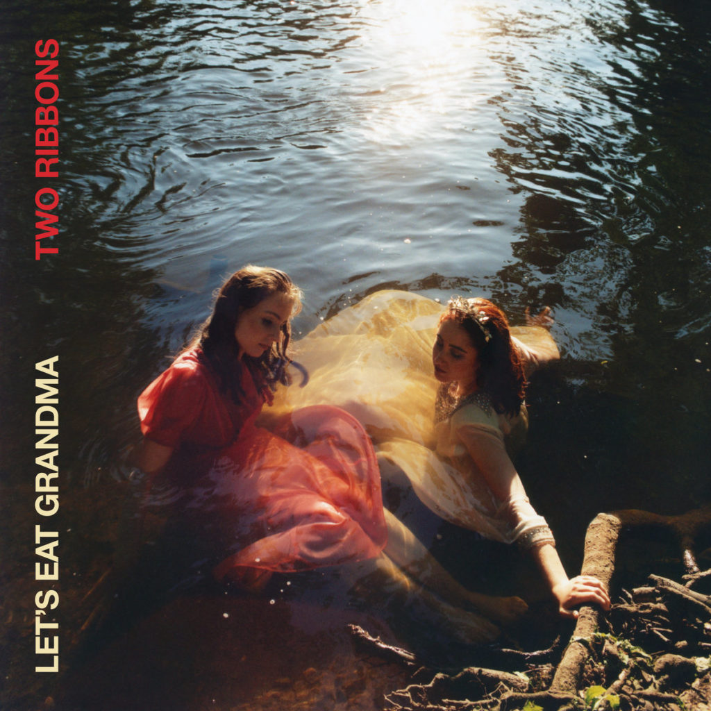 Zwei in Abendlicht getauchte Frauen in bunten Kleidern sitzen bis zu den Knien in einem ruhigen Fluss, umgeben von Uferwurzeln – Cover von „Two Ribbons“ von Let’s Eat Grandma.