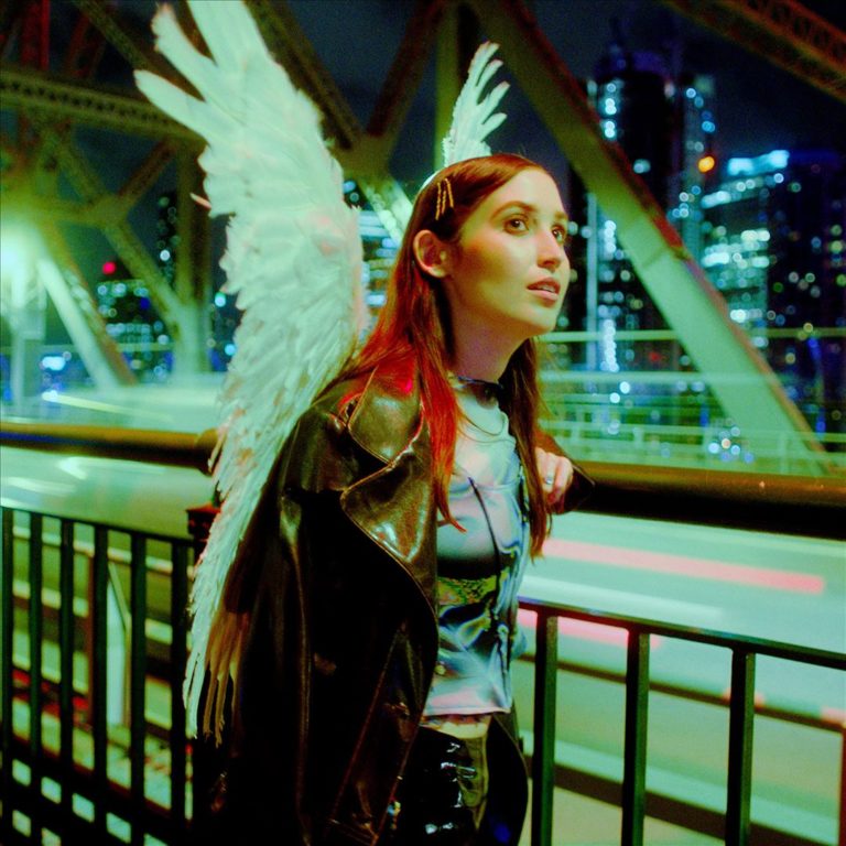 Hatchie steht auf einer nächtlichen Brücke, trägt Engelsflügel, im Hintergrund glitzern Stadtlichter und vorbeirauschender Verkehr.