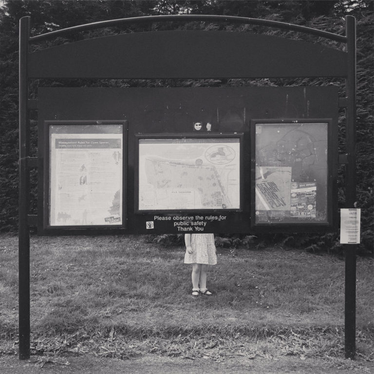 Schwarz-weißes Foto einer Park-Info-Tafel mit dem Hinweis „Please observe the rules for public safety“, hinter der nur die Beine eines Mädchens im Kleid auf einer Wiese zu sehen sind.