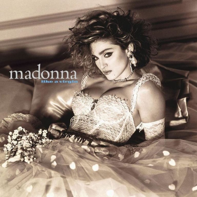 Madonna posiert in einem Spitzenkleid auf einem Bett, umgeben von Blumen.