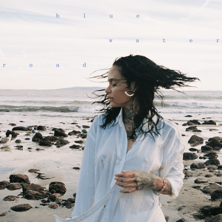 Kehlani steht am felsigen Strand, trägt ein weites helles Hemd, ihr Haar weht im Wind, während sie in die Ferne blickt.