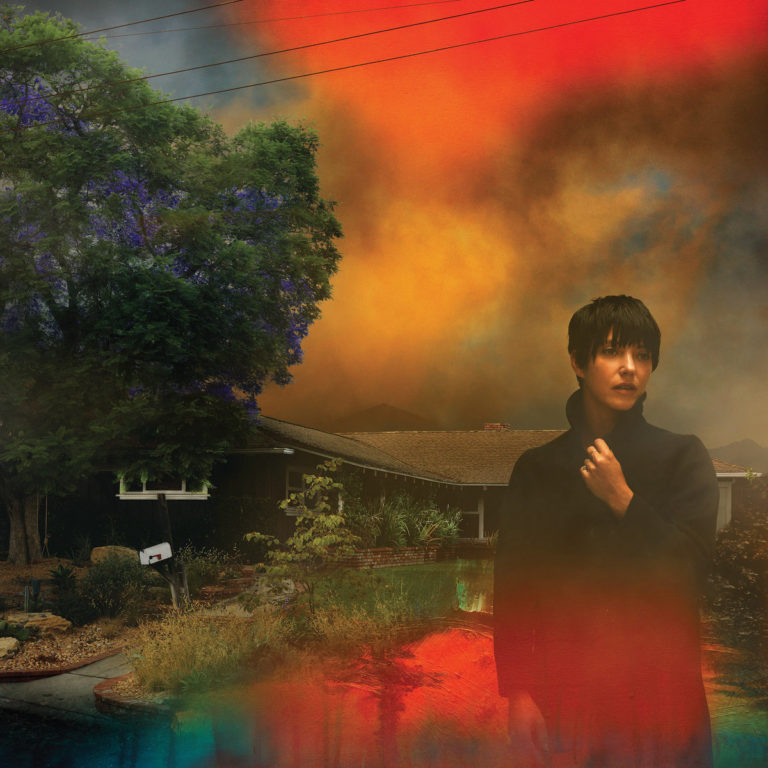 Sharon Van Etten steht in dunklem Mantel vor einem Vorstadthaus, während ein feuriges Lichtspiel den Himmel in Flammen taucht – Cover zu „We’ve Been Going About This All Wrong“.