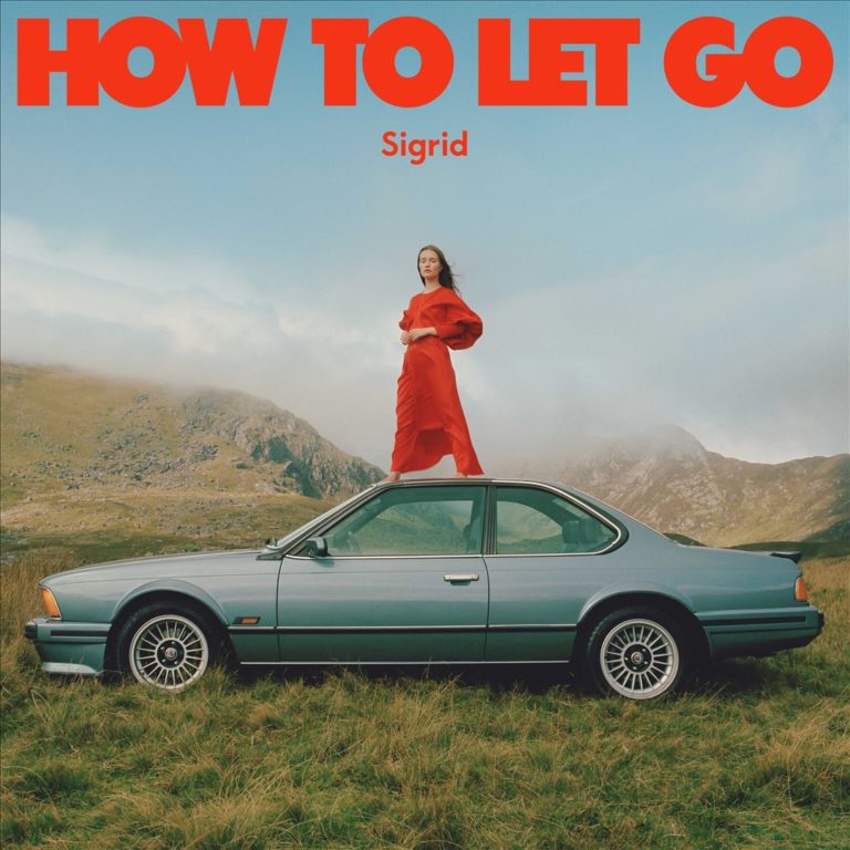 Frau in knallrotem Kleid steht auf dem Dach eines alten BMW inmitten nebliger Bergwiesen, darüber leuchtet in Großbuchstaben „HOW TO LET GO“ und „Sigrid“.
