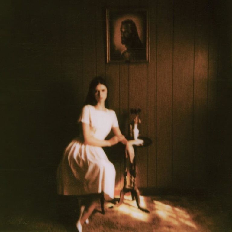Verschwommenes, düsteres Foto einer jungen Frau in weißem Kleid im schummrigen Raum mit Jesus-Porträt – Cover von Ethel Cains Debüt „Preacher’s Daughter“.