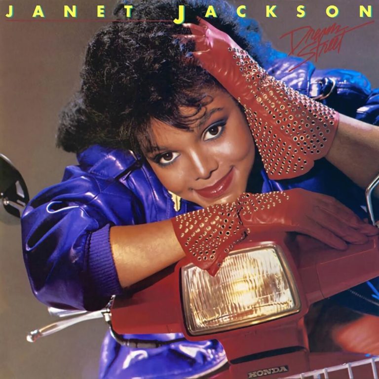 Janet Jackson posiert lächelnd mit einem roten Roller und trägt einen blauen Anzug sowie rote Handschuhe.