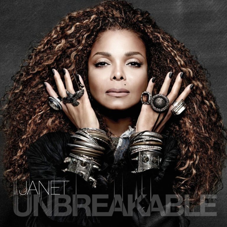 Janet Jackson mit lockigem Haar und Schmuck, frontal vor dunklem Hintergrund.
