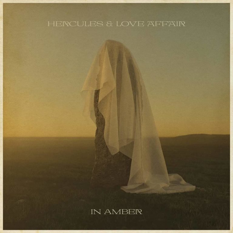Ein mystischer Monolith auf weiter Wiese, von einem transparenten weißen Schleier bedeckt – im goldenen Dämmerlicht eingefangen. Das Albumcover von „In Amber“ wirkt wie ein stilles Denkmal für Vergänglichkeit, Trauer und Transformation.