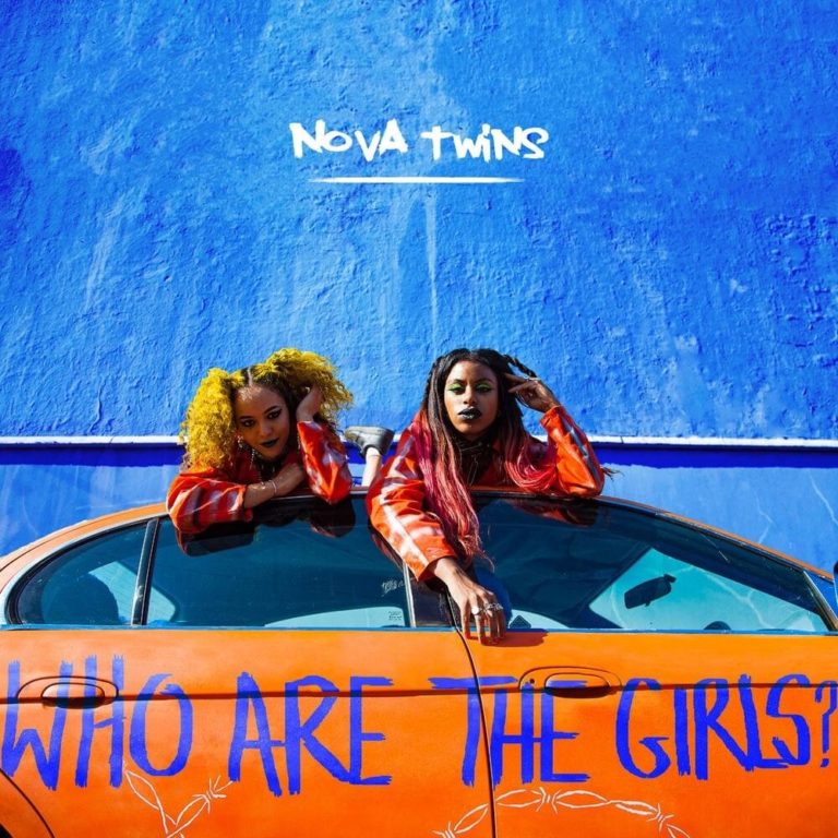 Zwei Frauen lehnen auf einem orangefarbenen Auto vor einer blauen Wand mit dem Schriftzug Nova Twins.