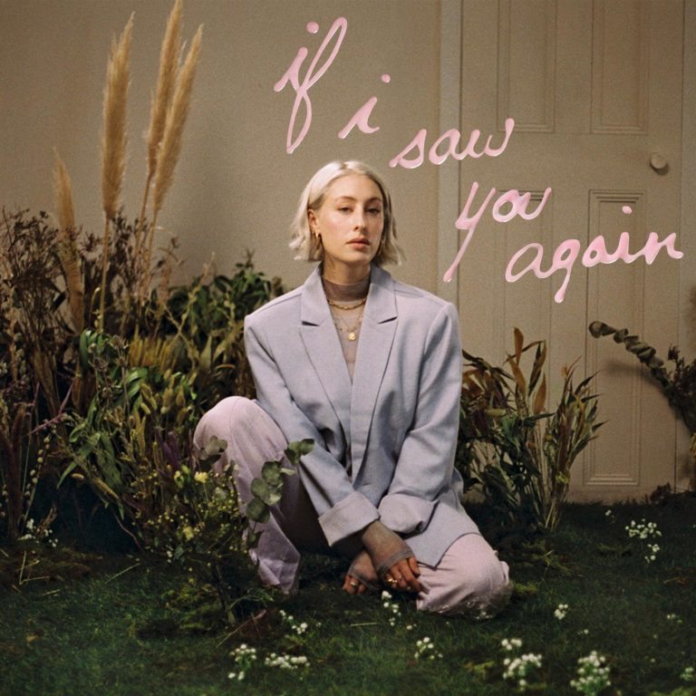 Albumcover von If I Saw You Again: Violet Skies sitzt in einem pastellfarbenen Anzug in einem Zimmer voller Pflanzen auf einem Teppich aus Moos. Über ihr ist der Albumtitel in rosa Handschrift an die Wand geschrieben – eine Szene zwischen Wohnzimmer und Traumbild, zwischen Leben und Erinnerung.
