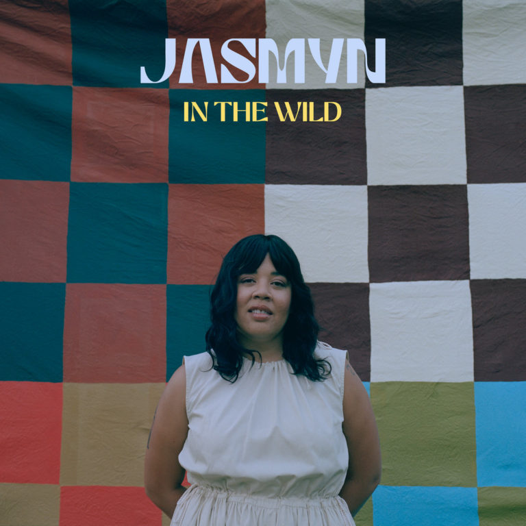 Albumcover von In The Wild zeigt Jasmyn vor einer farbenfrohen, karierten Decke – sie steht ruhig in einem cremefarbenen Kleid, mit offenem Blick, als würde sie gerade frisch in ihre neue kreative Freiheit treten.