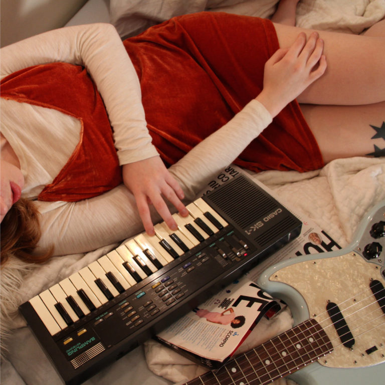 Person in rotem Kleid spielt Keyboard auf Bett neben Gitarre.