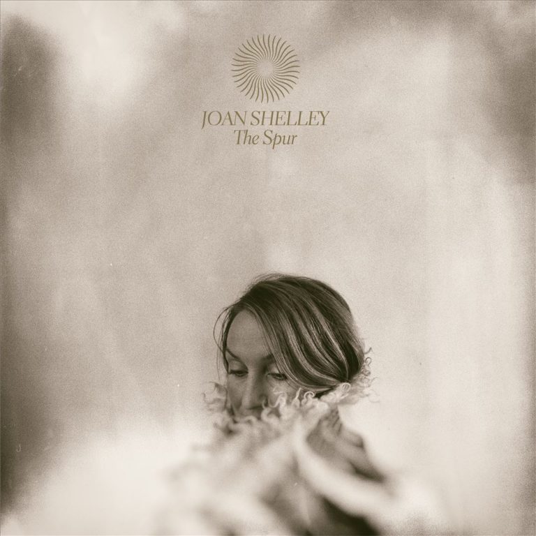 Cover des Albums „The Spur“ von Joan Shelley: In sepiafarbener Weichzeichnung ist nur ihr Gesicht sichtbar, halb von lockigem Stoff verdeckt, der ins Unschärfefeld gleitet. Oben zentriert in Goldschrift: Titel und Name.