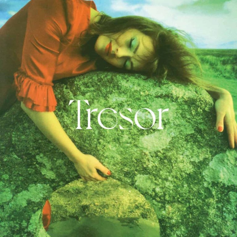 Das Cover von Gwenno Saunders’ „Tresor“ zeigt sie in rotem Kleid, wie sie verträumt an einen alten, moosbewachsenen Steinkreis lehnt. Ihre Augen geschlossen, die Lippen rot, das Make-up pastellfarben – im Hintergrund eine satte, grüne Landschaft unter einem leicht bewölkten Himmel. Das Wort „Tresor“ steht zentriert in eleganter Serifenschrift.