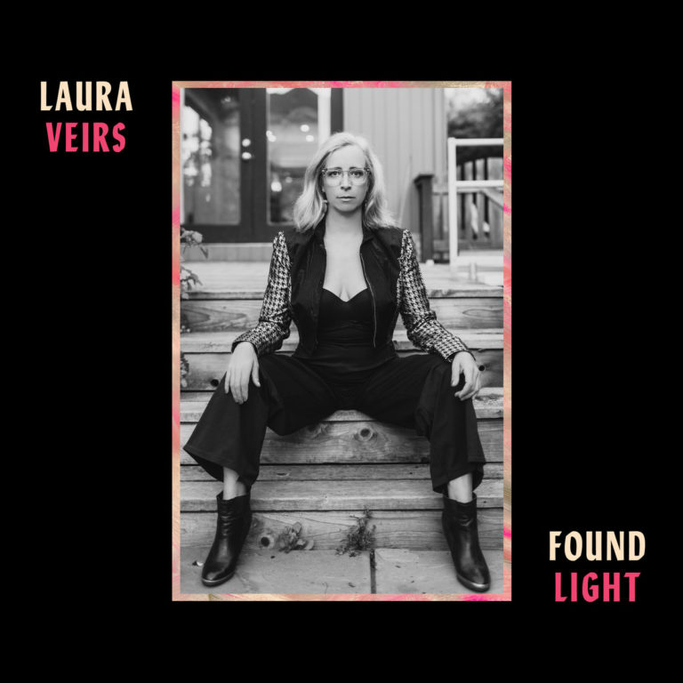 Schwarz-weißes Porträt der Musikerin Laura Veirs, die selbstbewusst auf Holzstufen sitzt, mit schwarzem Jumpsuit, karierter Jacke und klarer Haltung – eingerahmt von einem zarten, farbigen Rand, Titel und Name typografisch gesetzt auf schwarzem Hintergrund.
