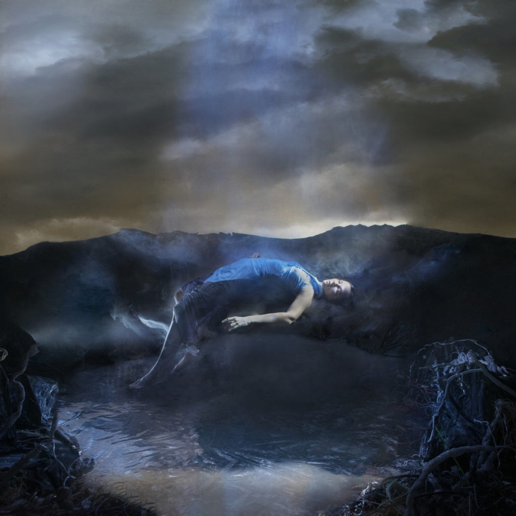 Caterina Barbieri liegt in trancehafter Haltung auf einer nebligen Erdlandschaft, von oben von blauem Licht durchdrungen. Das Artwork zu „Spirit Exit“ erinnert an eine entrückte Ophelia-Figur in digitaler Mystik – zwischen Kosmos, Natur und Abgrund.