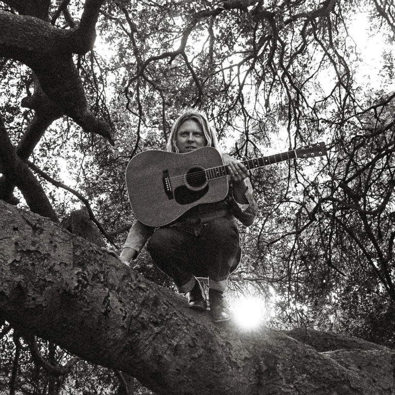 Ty Segall kauert mit Akustikgitarre auf einem knorrigen Ast im dichten Geäst – ein intimer Moment zwischen Natur, Schatten und Songwriting.