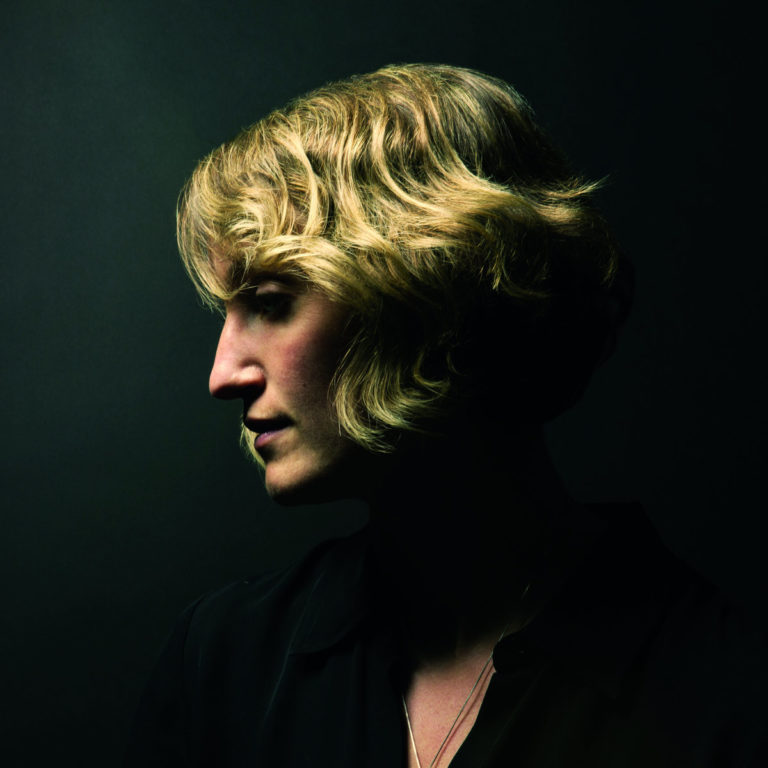 Profil einer Person mit welligem, blondem Haar vor dunklem Hintergrund