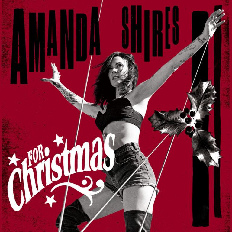 Amanda Shires posiert auf rotem Hintergrund mit Mistelzweig, Retro-Schriftzug „For Christmas“ und ausdrucksstarkem Bühnenlook – festlich und rebellisch.