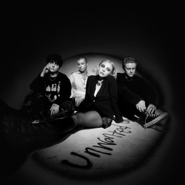 Schwarz-weißes Bandfoto als Cover von Unwanted von Pale Waves: Vierköpfige Band sitzt in einem dunklen, vignettartigen Lichtkreis, vor ihnen steht „Unwanted“ als Graffiti auf dem Boden geschrieben.