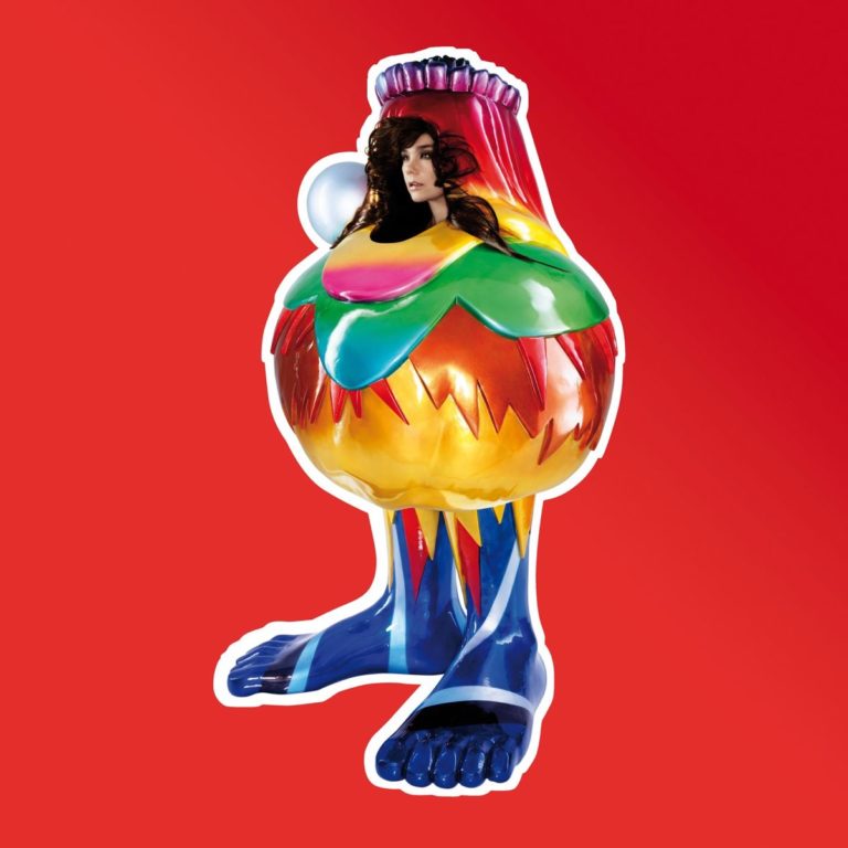 Bunte, skulpturartige Figur mit menschlichem Kopf auf rotem Hintergrund.