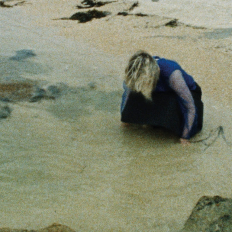 Eine blonde Person kauert in dunkler Kleidung an einem kargen Strand, den Blick gesenkt, die Hände im nassen Sand. Die Szene ist unscharf, analog anmutend und wirkt wie ein flüchtiger, intimer Moment – eingefangen zwischen Ebbe, innerem Rückzug und melancholischer Selbstbeobachtung. Ein visuelles Echo der verletzlichen Soundlandschaft von Elaine Howley.