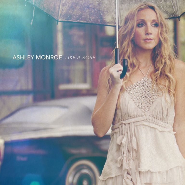 Ashley Monroe steht unter einem transparenten Regenschirm, trägt ein helles, perlenverziertes Kleid und blickt gedankenverloren zur Seite. Im Hintergrund ist ein unscharfes, dunkles Auto zu sehen. Links im Bild steht der Schriftzug „Ashley Monroe Like A Rose“.