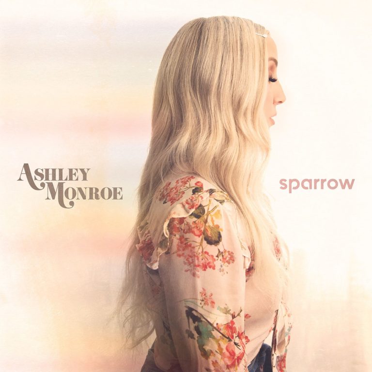 Seitliches Porträt von Ashley Monroe mit langen blonden Wellen und floral gemusterter Bluse, vor sanft verschwommenem, pastellfarbenem Hintergrund. Links der Schriftzug „Ashley Monroe“, rechts der Albumtitel „Sparrow“.