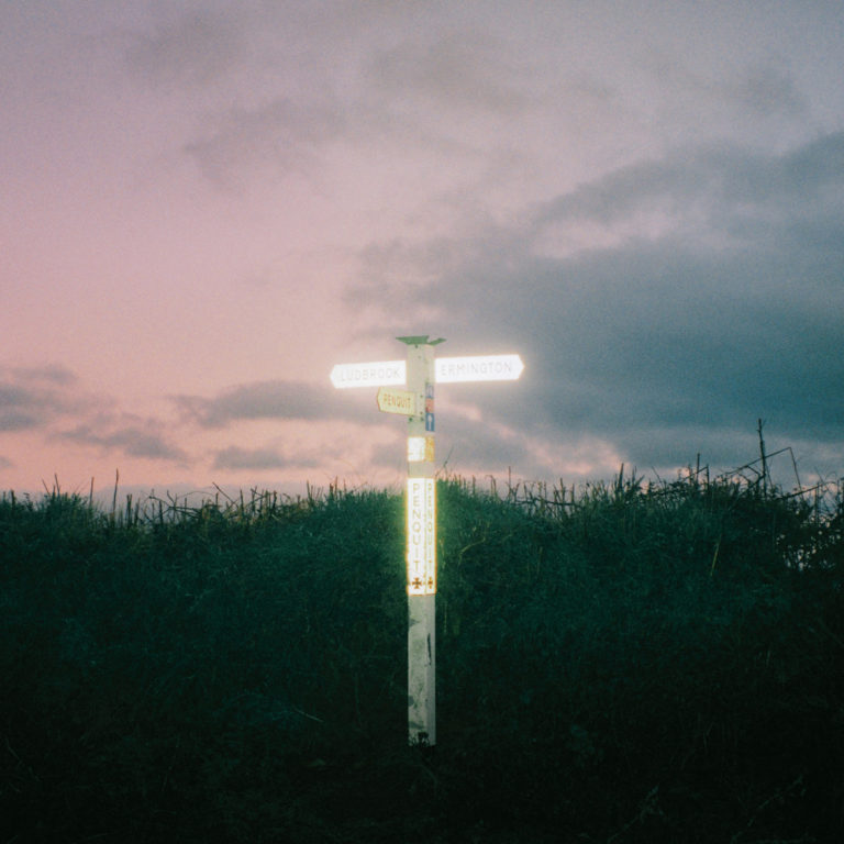 Das Cover zeigt ein beleuchtetes Wegweiserschild bei Dämmerung, umgeben von Hecken und lila-blauem Himmel – Artwork zum Album „Souvenirs“ von Pale Blue Eyes.