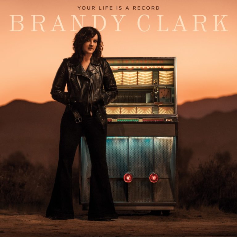 Frau in schwarzer Lederjacke steht neben einer Jukebox in einer Wüstenlandschaft bei Sonnenuntergang.