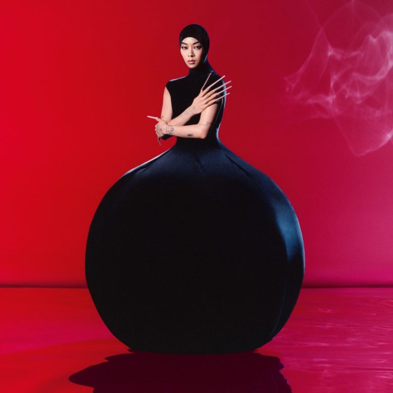 Rina Sawayama sitzt in einem überdimensionalen, skulpturalen schwarzen Kleid auf rotem Boden vor tiefrotem Hintergrund. Ihr Blick ist ernst, ihre Pose statuarisch. Ausdrucksstark, avantgardistisch und kontrolliert – wie ein Kunstwerk zwischen Modefotografie und Popikonografie.