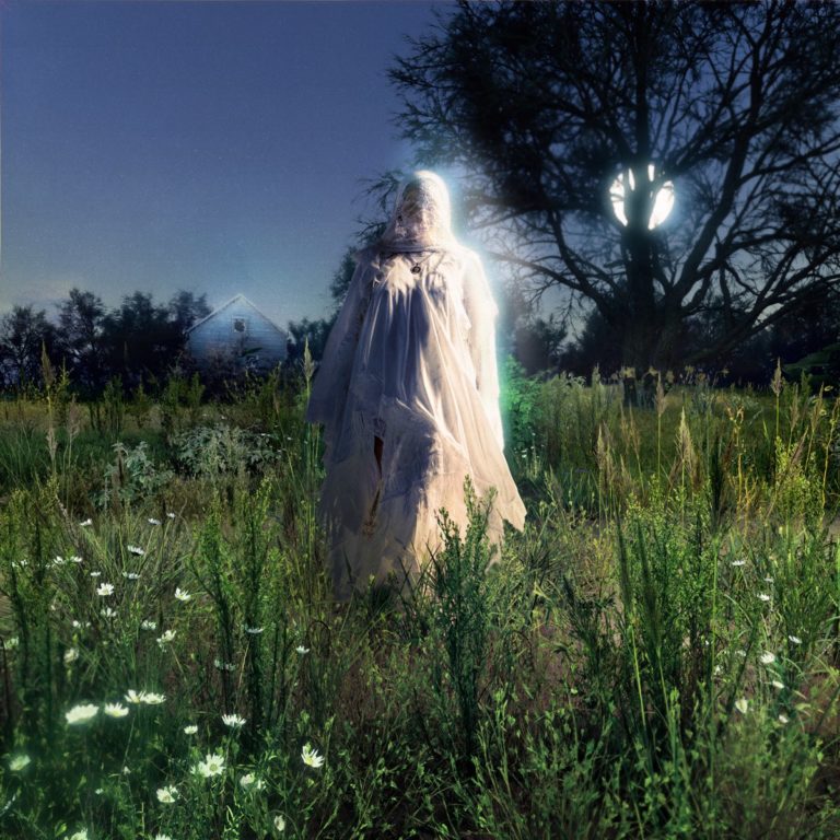 Eine in weißes Tuch gehüllte Figur steht nachts in einem überwucherten Feld, vom Mondlicht und einer geheimnisvollen Aura umgeben. Im Hintergrund ein kahler Baum mit Vollmond, daneben ein abgelegenes Haus – die Szene wirkt wie ein verträumter Geisterspaziergang zwischen Erinnerung und Aufbruch.